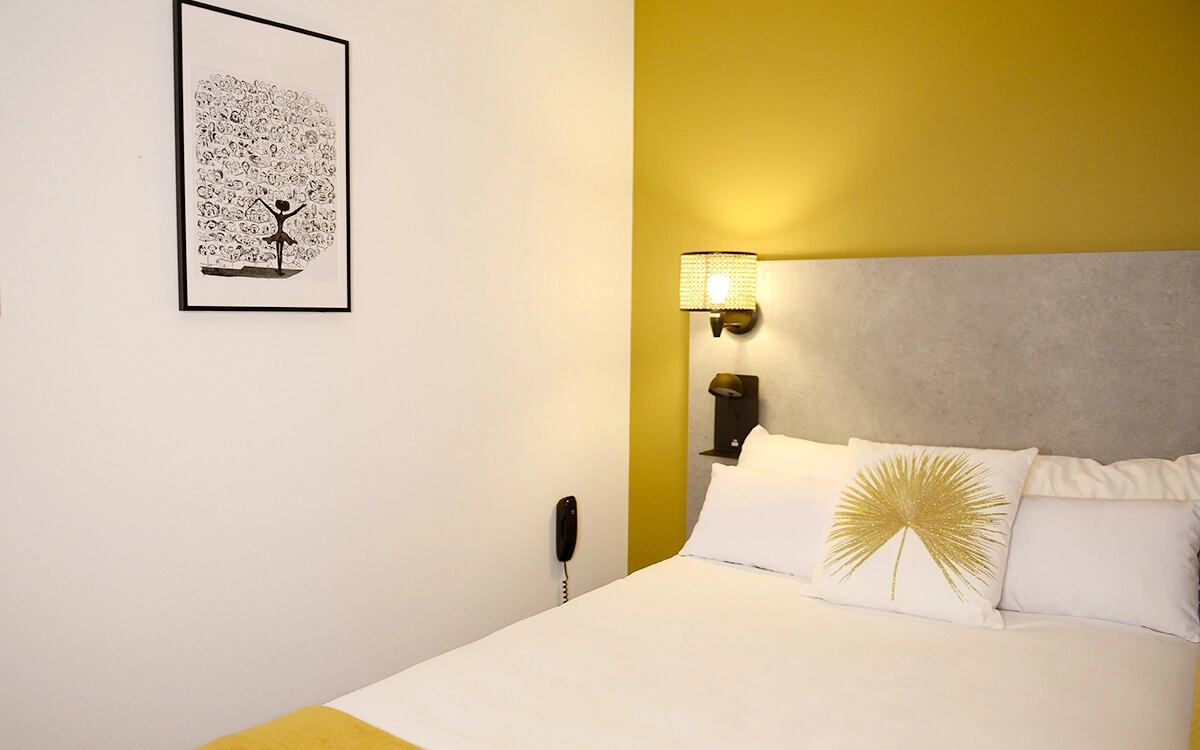 Chambre individuelle de l’Hôtel Villa Berlioz à Lyon, avec lit simple, tête de lit moderne, coussin décoratif et éclairage mural.